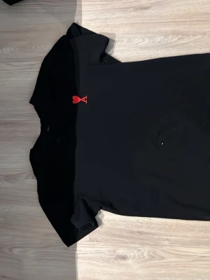 Svart t-shirt med rött hjärta Ami - Snygg svart t-shirt från AMI med ett litet rött hjärta och bokstaven A broderat på bröstet. Klassisk passform och rund halsringning. Perfekt för dig som gillar stilrena plagg med en twist.