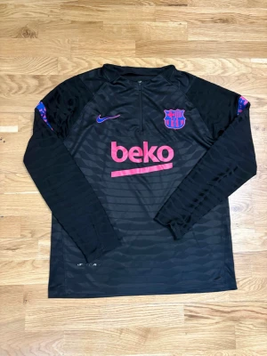 Svart FC Barcelona träningströja Nike - Svart långärmad träningströja från Nike med FC Barcelona-logga på bröstet och lila Nike Swoosh. Tröjan har halv dragkedja, rosa BEKO-tryck och diskret mönster i tyget. Perfekt för fotbollsträning eller sportiga dagar.