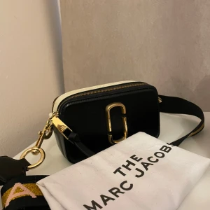 Marc Jacobs Snapshot väska  - Handväska från Marc Jacobs i modellen Snapshot. Väskan är svart med vita detaljer och har guldfärgade metalldetaljer. Axelremmen är bred och dekorerad med mönster i svart, rosa och gult. Lite små defekter såsom repor, men inget särskilt. Säljer då den inte kommer till användning. 