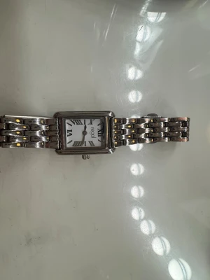 Silverfärgad klocka från Regal - Stilren klocka med silverfärgad länkarmband i metall och fyrkantig vit urtavla. Urtavlan har svarta romerska siffror och minimalistiska visare. Klassisk design som passar till många olika stilar.