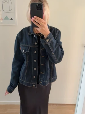 Mörkblå jeansjacka - Klassisk mörkblå jeansjacka med silverfärgade knappar och kontrastsömmar. Jackan har bröstfickor med lock och knapp samt en rak passform. Perfekt att slänga över vilken outfit som helst för en cool och tidlös look.