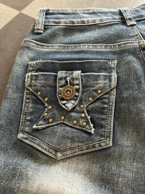 Blå bootcut jeans med stjärndetaljer - Säljer ett par blå bootcut jeans med slitningar och coola stjärndetaljer i nitar på bakfickorna. Jeansen har en knapp med text och kontrastsömmar som ger en unik look. Perfekta för dig som vill sticka ut med din stil!