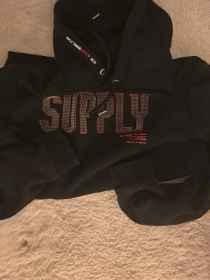 Svart hoodie från SUPPLY demand - Svart hoodie från SUPPLY med stort rött och vitt tryck på bröstet. Huva med dragsko och textdetaljer på snöret. Flera mindre tryck på ryggen. Skön och avslappnad passform, perfekt till streetwear-stilen.