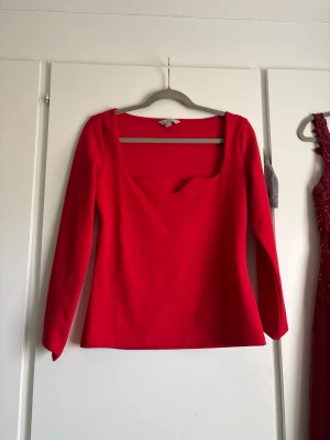 Röd långärmad topp med fyrkantig ringning - Snygg röd topp med lång ärm och fyrkantig ringning. Modellen är figurnära och enkel utan mönster eller detaljer. Perfekt att styla med jeans eller kjol för en trendig look.