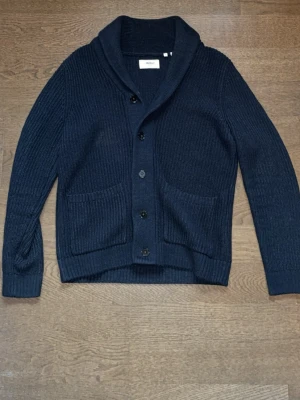 Mörkblå kofta från Norse Projects - Jävligt schysst cardigan med riktigt bra passform Snygg mörkblå kofta från Norse Projects med sjalkrage och knäppning framtill. Koftan är ribbstickad och har två stora fickor fram. Perfekt för lager på lager och ger en clean look till din outfit.