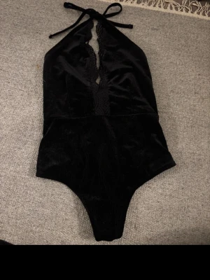 Svart halterneck body med spets - Säljer en svart body med halterneck och knytband i nacken. Bodyn har en djup v-ringning med dekorativ spetsdetalj framtill och är tillverkad i ett mjukt, sammetliknande material. Praktiska tryckknappar i grenen för enkel användning.öppen i ryggen