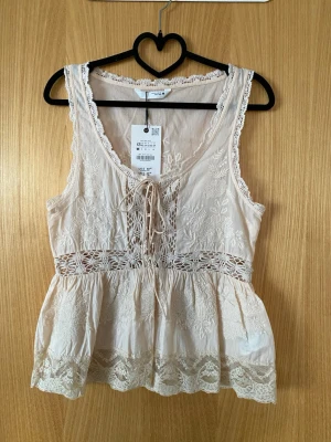 Beige broderad topp från Kiss the Sky - Söt beige topp från Kiss the Sky med broderade blommor, spetsdetaljer och knytning fram. Toppen är ärmlös, har v-ringning och dekorativ spets längs nederkanten och midjan. Perfekt för dig som gillar romantiska och ljusa plagg.