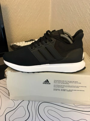 Svarta Adidas sneakers med mesh - Snygga svarta Adidas sneakers med mesh-överdel och klassiska tre ränder på sidan. Skorna har en vit sula och svarta snören, samt en stabil hälkappa. Perfekt för dig som gillar stilrena och sportiga skor.