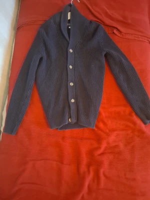 Mörkblå kofta med sjalkrage - Mörkblå ribbstickad kofta från It's So Simple To Dress Up. Koftan har sjalkrage, fem knappar framtill och långa ärmar. Perfekt för lager på lager och ger en clean look till din outfit.