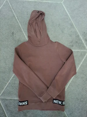 Brunröd hoodie med tryck och huva - Säljer en brunröd hoodie med huva och svartvita textdetaljer längst ner där det står 'NEW. VIS #NS'. På ryggen finns ett stort svart tryck med olika bokstäver och siffror. Hoodien har långa ärmar och ribbade muddar. Perfekt för en avslappnad streetwear-look.