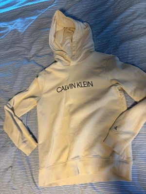 Vit hoodie från Calvin Klein - Snygg vit hoodie från Calvin Klein med stor logga på bröstet och liten 'JEANS'-patch bak. Klassisk huva och ribbade muddar vid ärmar och nederkant. Tillverkad i mjukt bomullsmaterial, perfekt för en clean och stilren look.