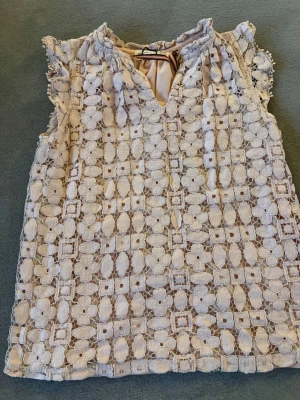 Vit spetsblus med volangdetaljer - Supersöt vit blus med broderad spets och volangdetaljer vid axlarna. Blusen har v-ringning framtill och knäppning bak i nacken. Perfekt för dig som gillar romantisk och feminin stil.