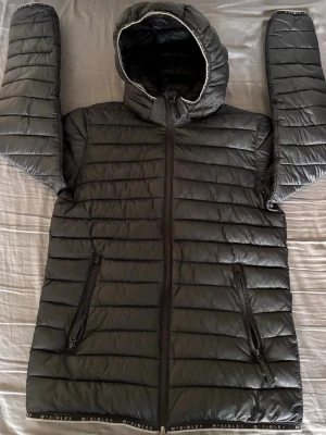 Svart dunjacka med huva från McKinley - Säljer en svart dunjacka från McKinley med huva och två dragkedjeförsedda fickor. Jackan har quiltad design och är lätt vadderad, perfekt för kalla vinterdagar. Diskret logotyp på insidan och längs nederkanten. Stängs med dragkedja framtill. Storlek: 158/164(som XS)