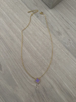 Guldhalsband med lila fyrklöverhänge - Snyggt halsband i guldfärgad metall med ett lila fyrklöverformat hänge. Kedjan är tunn och elegant, och hänget har små guldfärgade detaljer runt kanterna. Perfekt för dig som gillar färg och unika smycken.