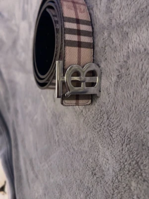 Burrberry bälte  - Snyggt bälte med klassiskt rutmönster i beige, svart och brunt på ena sidan och mörkgrått rutmönster på andra sidan. Bältet har ett stilrent silverfärgat spänne med unik design. Perfekt accessoar för att lyfta din outfit.