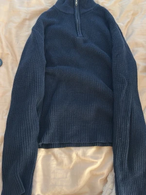 Mörkblå stickad halvzip från 157 - Mörkblå stickad tröja från 157 med halvzip och hög krage. Tröjan har ribbstickad struktur, långa ärmar och en avslappnad passform. Perfekt för lager på lager och enkel att styla till olika outfits.