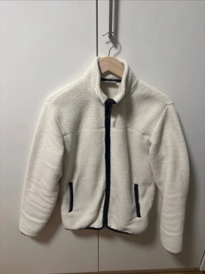 vit/beige fleecekofta med svarta drag - Mysig vit teddyjacka med hög krage och dragkedja framtill. Jackan har svarta detaljer längs dragkedjan och vid fickorna, samt två sidofickor. Perfekt för kalla dagar och ger en riktigt chill vibe. storlek Xs men funkar på S och M