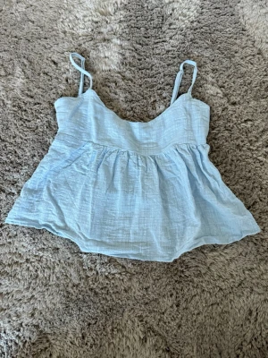 Ljusblå linnetopp med knyt i ryggen - Supersöt ljusblå linnetopp med tunna axelband och volangliknande nederdel. Toppen har en söt knytning i ryggen som ger en extra detalj. Perfekt för varma dagar och enkel att styla till jeans, kjol eller linnebyxor.