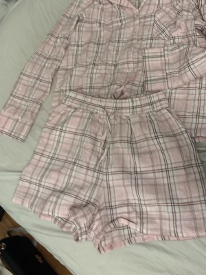 Rosa rutigt pyjamasset med shorts - Sött pyjamasset i ljusrosa med rutigt mönster i vitt och grått. Setet består av en långärmad skjorta med knappar och bröstficka samt matchande shorts med resår i midjan. Perfekt för mysiga kvällar hemma.
