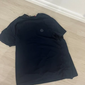 Svart t-shirt från Moncler - Säljer en svart t-shirt från Moncler med diskret logga på bröstet. Klassisk passform och rund halsringning. Perfekt till jeans eller shorts för en clean och enkel stil.