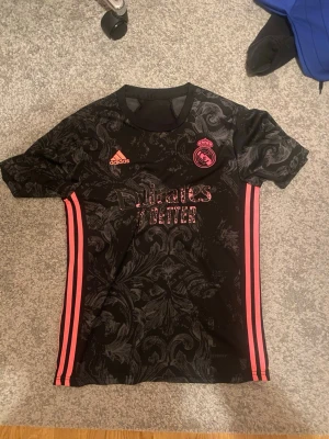 Real Madrid svart tränings t-shirt - Snygg svart Real Madrid tränings t-shirt från Adidas med rosa detaljer och klubbmärke på bröstet. Tröjan har ett mönstrat tyg, korta ärmar och rosa ränder längs sidorna. Perfekt för fotbollsträning eller gymmet.