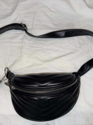 Svart magväska från Bershka - Snygg svart mag/axel väska från Bershka i slätt, glansigt material som ger en modern look. Väskan har en enkel, stilren design med justerbar axelrem och passar perfekt till alla outfits. Perfekt för dig som gillar minimalistisk stil.