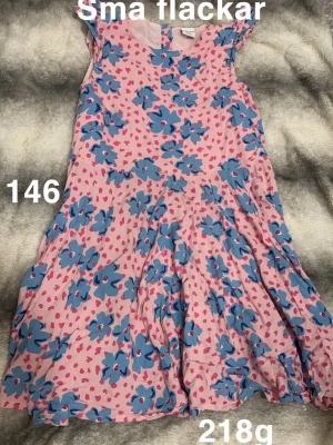 Rosa klänning med blå blommor - Söt rosa klänning med stora blå blommor och små rosa hjärtan som mönster. Klänningen har kort ärm och en lätt utställd kjol. Perfekt för dig som gillar färgglada och lekfulla plagg.🌸 Har mindre fläckar. Storlek 146