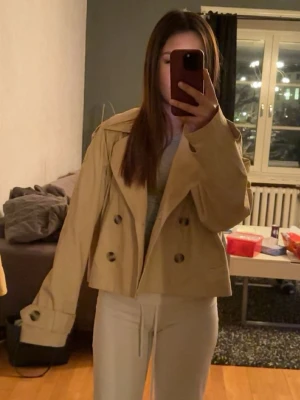 Kort beige trenchcoat från H&M - Snygg kort trenchcoat från H&M i beige bomullsmix. Jackan har dubbelknäppning med stora knappar, bred krage och axelklaffar. Ärmarna har dekorativa remmar vid mudden och jackan har en boxig, trendig passform. Perfekt för lager på lager och streetstyle.