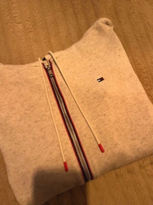 Beige hoodie från Tommy Hilfiger - Säljer en beige hoodie från Tommy Hilfiger med dragkedja och röda detaljer på snören och dragkedja. Klassisk logga på bröstet och mjukt material. Perfekt för dig som gillar stilrena och bekväma plagg.