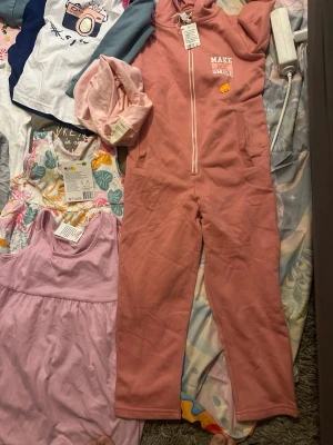 Rosa jumpsuit med huva och dragkedja - Mysig rosa loungewear jumpsuit med huva och vit dragkedja framtill. Trycket 'MAKE ME SMILE' på bröstet och två fickor i sidorna. Perfekt för chill dagar hemma. Mjuk och skön bomullsblandning.