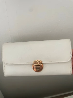 Vit plånbok med gulddetalj - Stilren vit plånbok i konstläder med snygg guldfärgad vridknäppe framtill. Insidan har flera kortfack och ett separat fack med dragkedja för mynt. Perfekt för dig som gillar enkel och klassisk design.