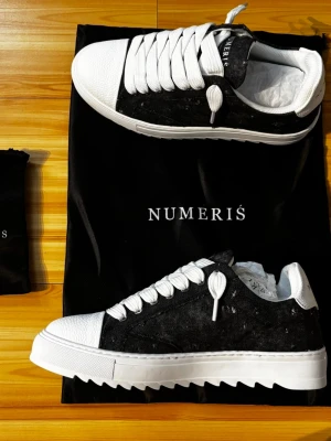 Svarta och vita sneakers från Numeris - Snygga sneakers från Numeris med svart ovandel och vita detaljer. Skorna har breda vita snören, vit tå och häl samt en unik såldesign med räfflad kant. Materialet ser ut att vara canvas och läder. Perfekt för dig som vill sticka ut med din stil.