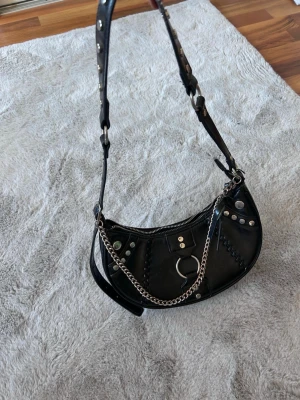 Svart axelväska med kedjedetaljer - Cool svart handväska i halvmåneform med blanka ytor, silverfärgade detaljer och kedja framtill. Väskan har dragkedja upptill och ett litet fack inuti med dragkedja. 