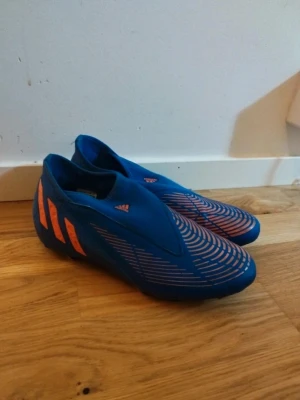 Blå Adidas fotbollsskor utan snörning - Säljer ett par blå fotbollsskor från Adidas med orangea detaljer och ikoniska stripes på sidan. Skorna har en modern design utan snörning och mönstrad ovandel för bättre bollkontroll. Perfekta för dig som vill sticka ut på planen och ha grym touch.
