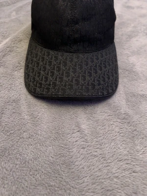 Svart Dior keps med monogram - Snygg svart keps från Christian Dior med klassiskt Dior-monogram över hela kepsen och böjd skärm. Justerbar rem bak i svart läder med präglad logga. Perfekt accessoar för en trendig streetwear-look.