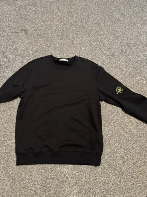 Svart sweatshirt från Stone Island - Svart crewneck sweatshirt från Stone Island med den klassiska logotypen på vänster ärm. Tröjan har rund halsringning, ribbade muddar och är tillverkad i mjukt bomullsmaterial. Perfekt för en clean och stilren look.