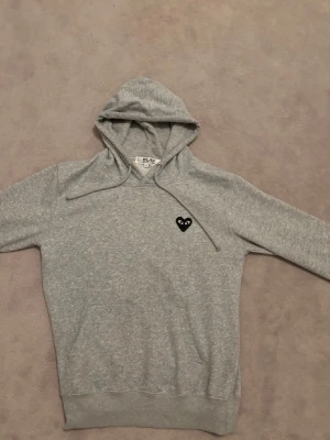 Grå Comme des Garçons PLAY hoodie - Snygg grå hoodie från Comme des Garçons PLAY med svart hjärta och ögon på bröstet samt stort svart hjärta med ögon på ryggen. Klassisk huva med dragsko och känguruficka framtill. Tillverkad i mjuk bomullsblandning, perfekt för en chill och trendig look.