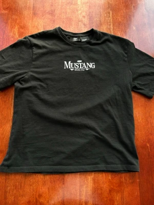 Svart Mustang Boss 302 t-shirt House - Svart t-shirt från House med Mustang Boss 302-tryck framtill. Klassisk rund hals och korta ärmar. Tillverkad i mjuk bomull och officiellt licensierad av Ford. Perfekt för dig som gillar bilar och vill ha en stilren look.