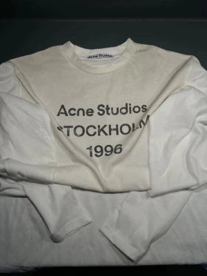 Vit långärmad topp Acne Studios - Vit långärmad topp från Acne Studios med tryck 'Acne Studios STOCKHOLM 1996' på bröstet. Klassisk rund halsringning och relaxed fit. Perfekt för dig som gillar minimalistisk och stilren design.
