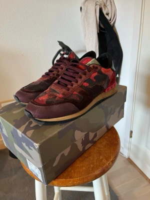 Valentino röda camo sneakers - Snygga Valentino Rockrunners i röd och vinröd camo-design.  Materialmix av mocka och textil ger en exklusiv känsla. Perfekta för dig som vill sticka ut med en unik look. Storlek 42 men lite stora i storlek. Låda och dustbags ingår. Kan gå ner lite i pris vid snabb och smidig affär. 👌 bara att fråga om du undrar något 