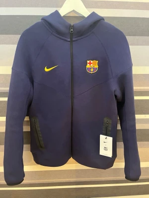 Nike FC Barcelona lila zip hoodie - Blå hoodie från Unisports med FC Barcelona-märke på bröstet och gul Nike-logga. Dragkedja framtill, två fickor med dragkedja och huva. Helt ny, inte använda någon gång nytt pris 1186 kr!