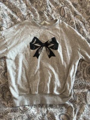 Grå tröja med svart rosett - Supersöt grå sweatshirt med en stor svart rosett framtill. Tröjan har rund halsringning, ribbade muddar och är gjord i mjukt sweatshirtmaterial. Perfekt för en chill och trendig look.