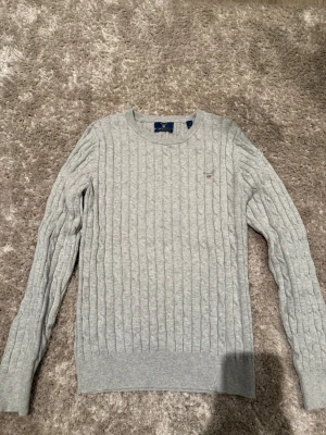 Ljusgrå kabelstickad tröja GANT - Säljer en ljusgrå kabelstickad tröja från GANT i stretchig bomull. Tröjan har rund halsringning, ribbade muddar och en liten GANT-logga broderad på bröstet. Perfekt för dig som gillar klassisk och stilren design.