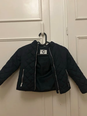 Mörkblå quiltad pufferjacka från Cubus - Stilren mörkblå pufferjacka från Cubus med quiltat mönster och silvriga dragkedjor. Jackan har rund halsringning och två fickor framtill. Perfekt för kalla dagar och ger en cool look till din outfit.