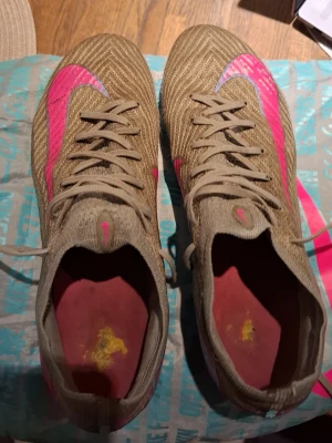 Nike Mercurial bruna fotbollsskor elite - Säljer ett par Nike Mercurial fotbollsskor i brunt med rosa detaljer och Nike-logga. Skorna har snörning, låg profil och ovandel i textilmaterial för bra bollkontroll. Snygg design med rosa sula och insida. Perfekta för dig som vill sticka ut på planen.