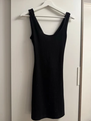 Svart ribbad ärmlös klänning - Svart ribbad klänning med tight passform och ärmlös design. Klänningen har en djup urringning fram och öppen rygg med cut-out detalj. Perfekt för dig som gillar minimalistisk och stilren look.