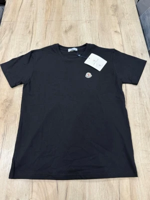 Svart Moncler t-shirt med logga - Snygg svart t-shirt från Moncler med klassisk rund hals och korta ärmar. På bröstet sitter Monclers ikoniska logga som en patch. Tillverkad i mjuk bomull för en skön känsla och enkel stil.