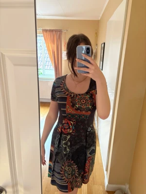 Mönstrad kortklänning med färgglada detaljer - Kortklänning med färgglada blommiga och randiga mönster i svart, rött, grönt och blått. Klänningen har rund halsringning och korta ärmar. Materialet känns mjukt och stretchigt, troligen bomull eller polyester. Perfekt för dig som gillar unika prints och bekväm passform. Fint skick.  Använd väldigt få gånger.  Inga fel på den som jag kan se. Storlek: M  Material: se sista bild  PRIS KAN ALLTID DISKUTERAS!