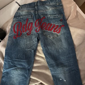 BDG Jeans med röd brodyr - Blå jeans från BDG med coolt röd broderad text 'Bdg Jeans' över bakfickorna. Jeansen har klassisk femficksdesign, raka ben och slitna detaljer samt vita färgstänk framtill. Perfekta för dig som vill sticka ut med en unik streetstyle. En liten hyska som är av,se bild. Nypris 650. Storlek w27 lenght 32