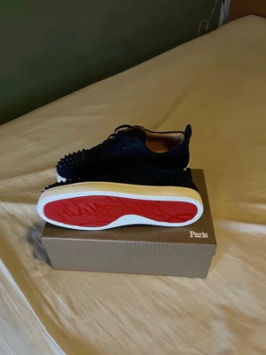 Mörkblå Louboutin sneakers med nitar - Snygga mörkblå sneakers från Christian Louboutin i mocka med svarta nitar på tån och klassisk röd sula. Skorna har vita sulor och svarta skosnören, perfekt för dig som vill sticka ut med en edgy och exklusiv look.